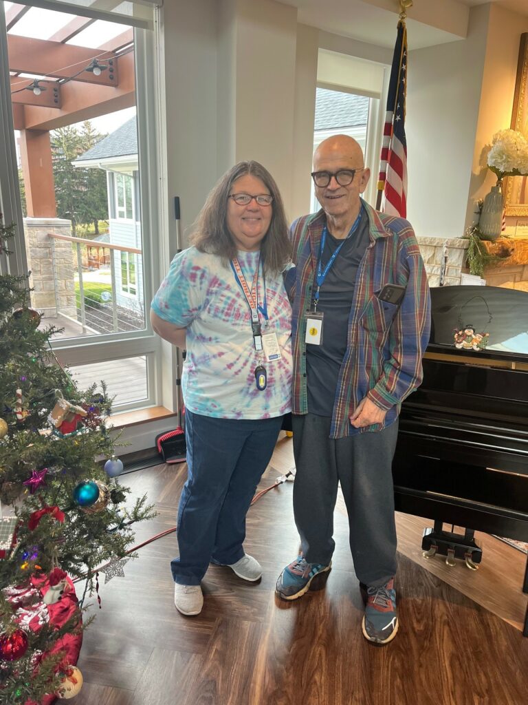 A man and woman stand between a Christmas tree, US flag, and piano.